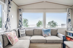 A cozy seating area with a sofa and cushions, large windows with a view, and a small table with a clock. - Cabin - Sleeps 4 - Pets Ok - Balcony - Parking (Watchet)