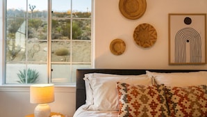 Un dormitorio con cama, lámpara de noche, ventana que ofrece vistas a un paisaje desértico, y decoración en las paredes que incluye platos tejidos y una obra de arte abstracta enmarcada.