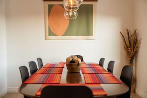Uma mesa de jantar com um arranjo central, rodeada por cadeiras, num quarto com uma obra de arte emoldurada na parede.