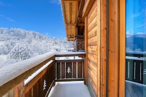 A wooden balcony with a snow-covered railing, a clear sky, and a snowy mountain landscape in the background.