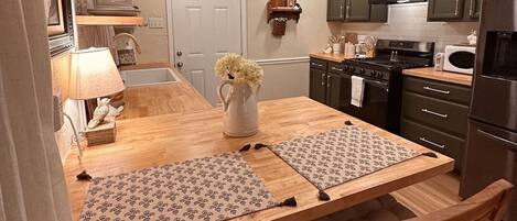 Eine kleine Küche mit dunklen Schränken, einer Mikrowelle, einem Backofen und einem Spülbecken. Ein Holztisch mit Untersetzern und einem Blumenarrangement steht daneben.