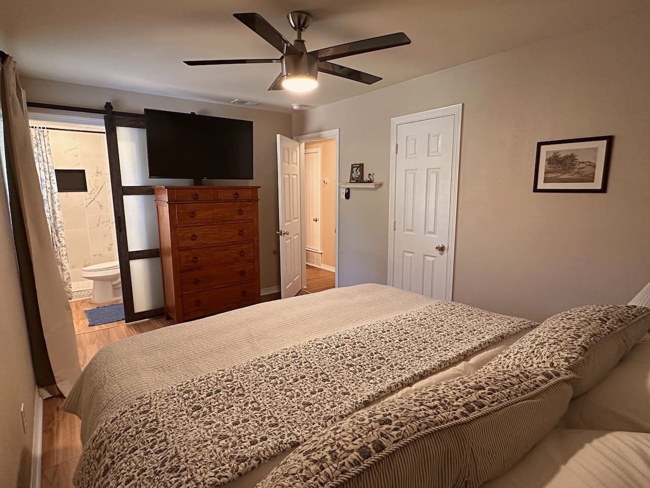 A bedroom with a bed, a ceiling fan, a dresser, a television, and a door leading to a bathroom.