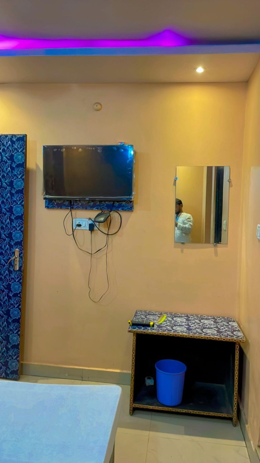 Une chambre à coucher avec un lit, un téléviseur fixé au mur, une petite table avec un miroir et une poubelle bleue.
