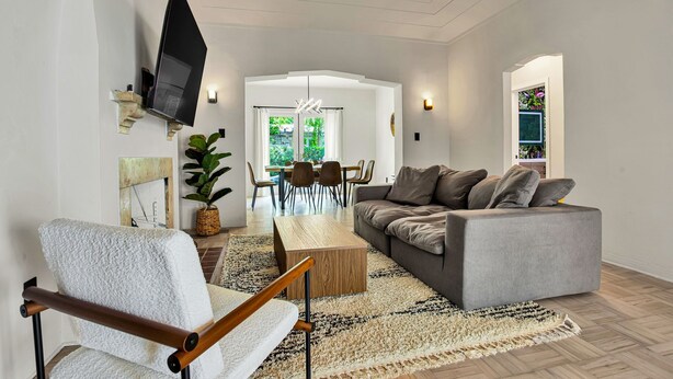 A modern living room with a grey sofa, a wooden coffee table, and a white armchair. There is a TV mounted on the wall, a dining area with chairs and a table, and a large window with a view of greenery. - Villa in the heart of design district! POOL+LOCATION (Miami)