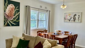 Een woonkamer met een beige bank, een eettafel met stoelen, een raam met gordijnen, en kunstwerken aan de muren.