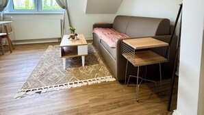 A modern living room with a sofa, a coffee table, a small table, and a lamp. There is a window with curtains, a rug, and a ceiling light. - Apartment at the Obersee (Bielefeld)