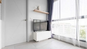 Un salon moderne avec un téléviseur à écran plat fixé sur un meuble blanc, une étagère en bois au-dessus et une grande fenêtre ornée de rideaux légers.