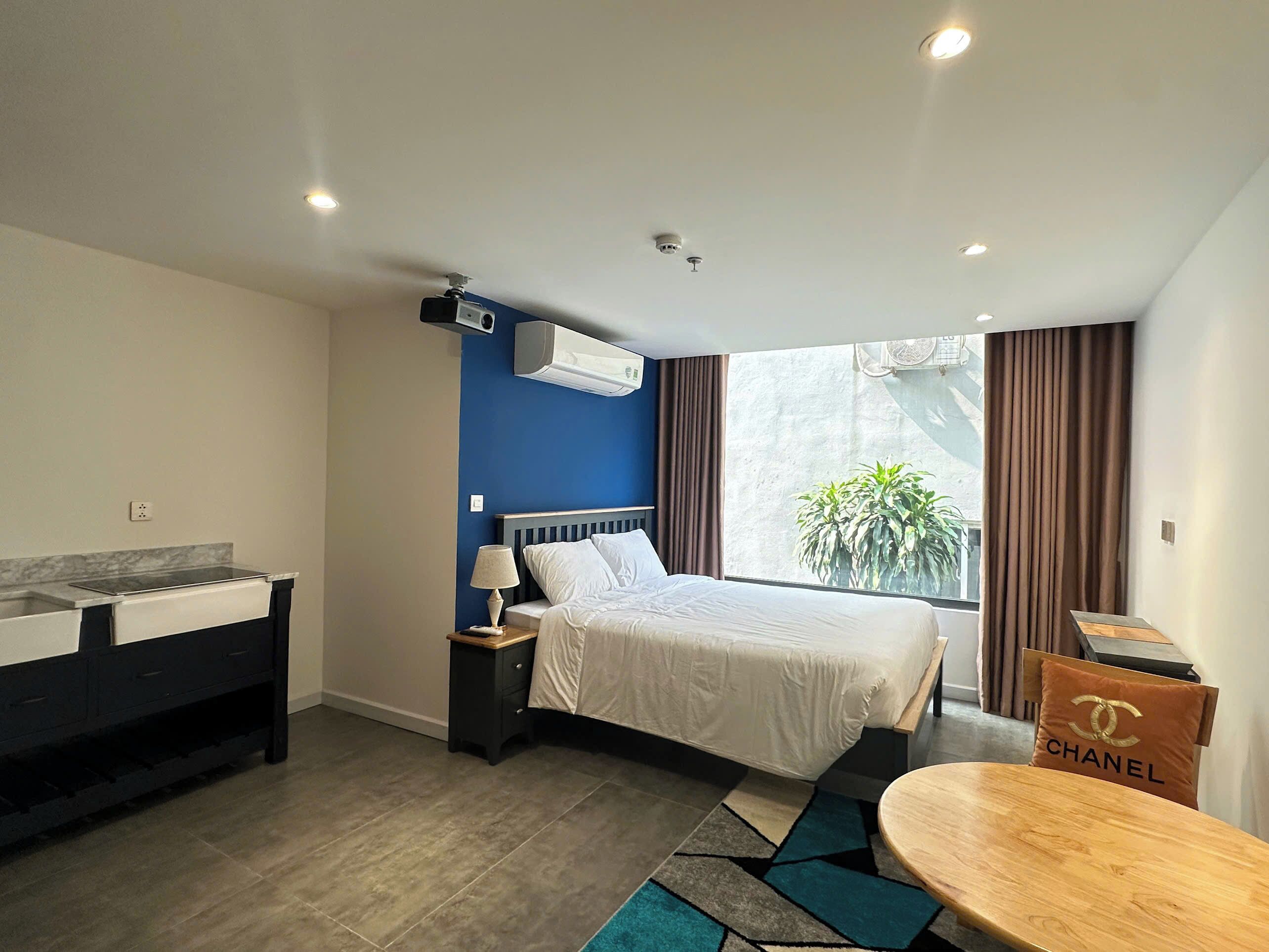 A modern hotel room with a bed, bedside table, lamp, air conditioning unit, and a view of the outdoors.