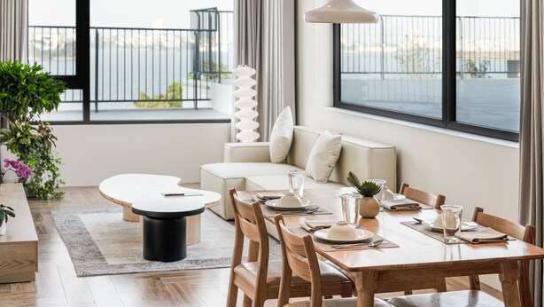 A modern dining area with a wooden table set for four, a white sofa, and a view of the water through large windows. - Bach Plateaus Hotel (Hanoi)