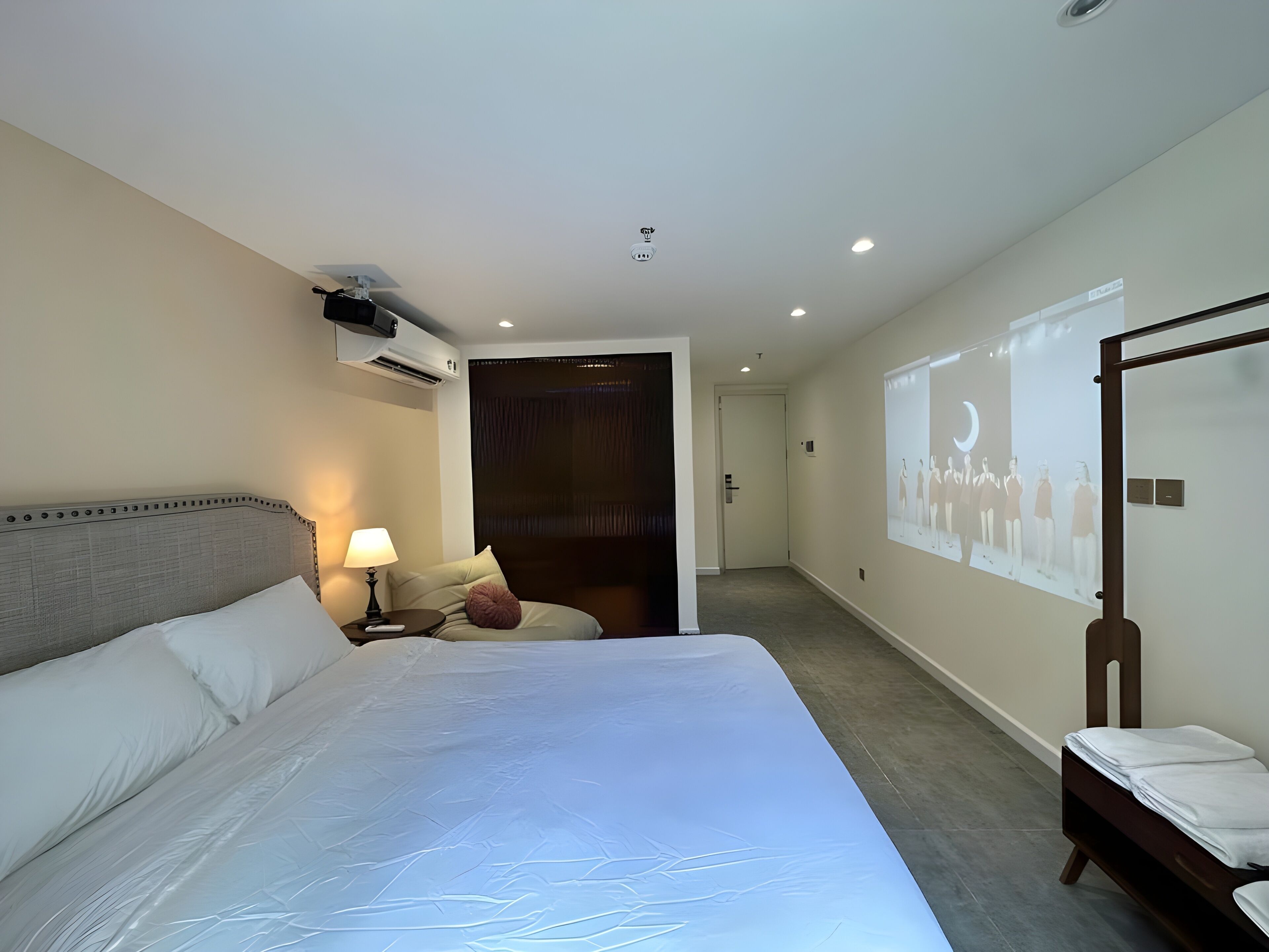 A hotel room with a large bed, a bedside table with a lamp, a wall-mounted air conditioner, and a projection screen displaying a group of people.