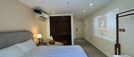 A hotel room with a large bed, a bedside table with a lamp, a wall-mounted air conditioner, and a projection screen displaying a group of people.