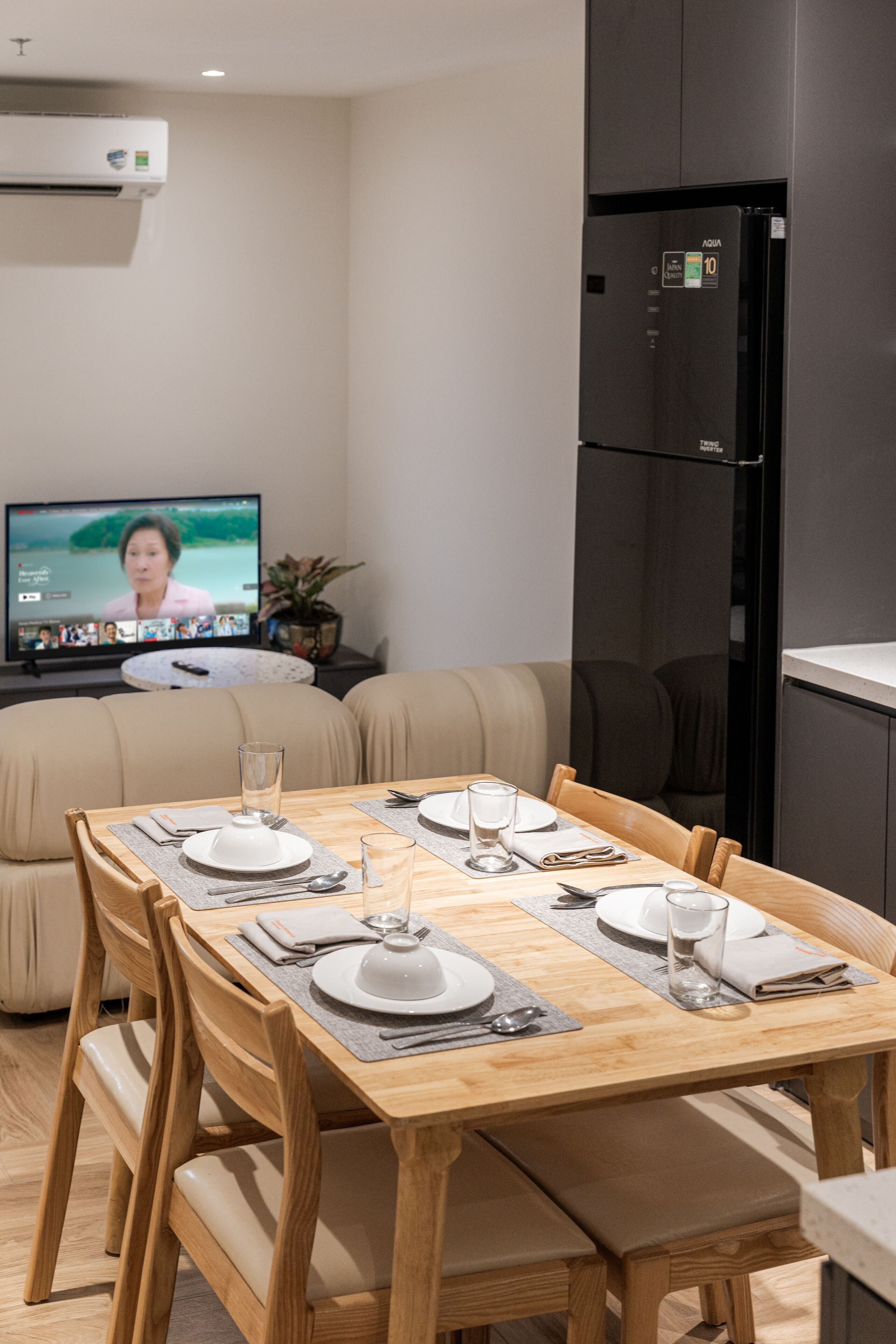 A modern living room with a dining area, a black refrigerator, a flat-screen TV, and a beige sofa.