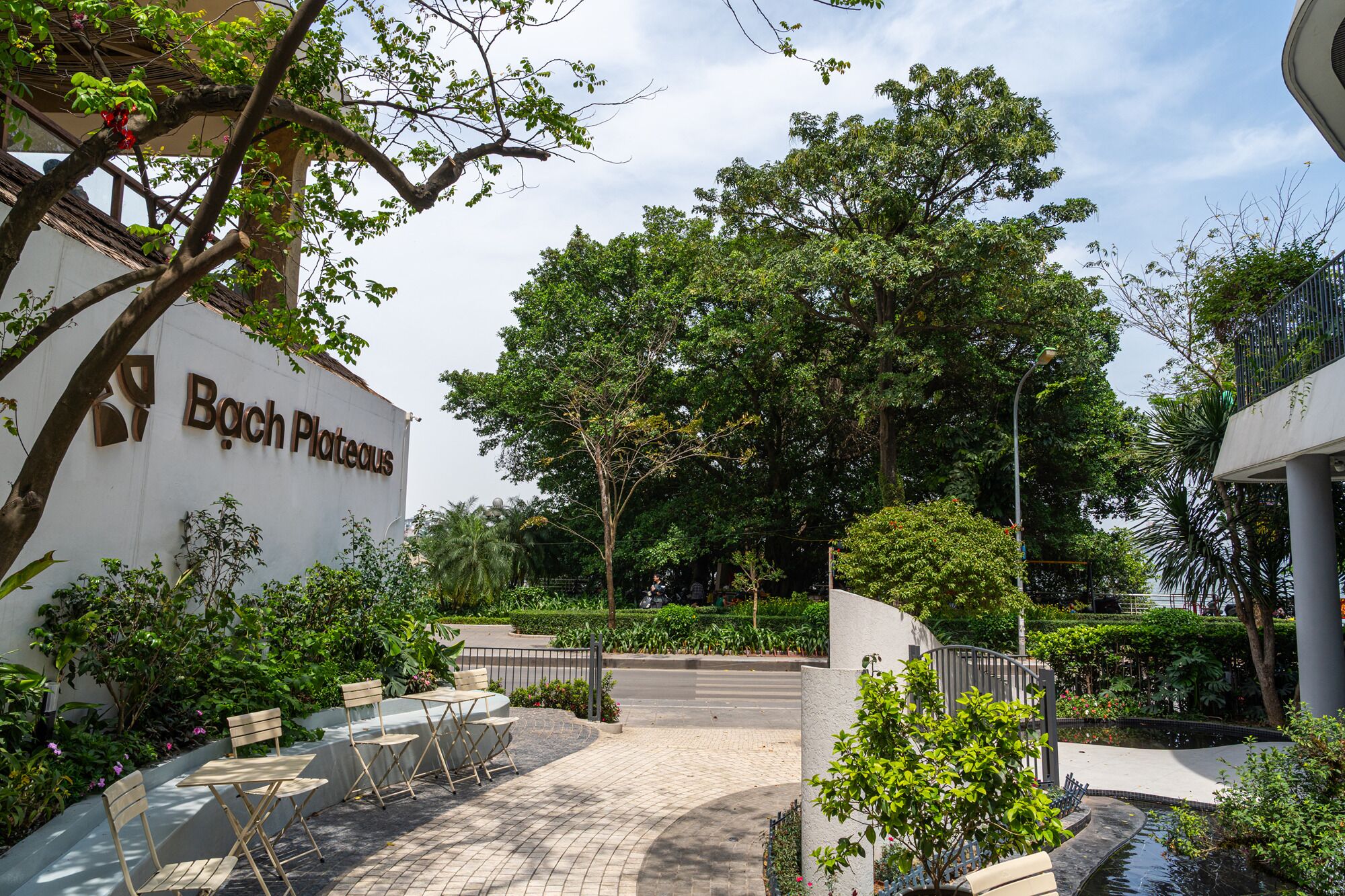 A courtyard with a sign reading "Bach Plateaus," surrounded by lush greenery and a small water feature.