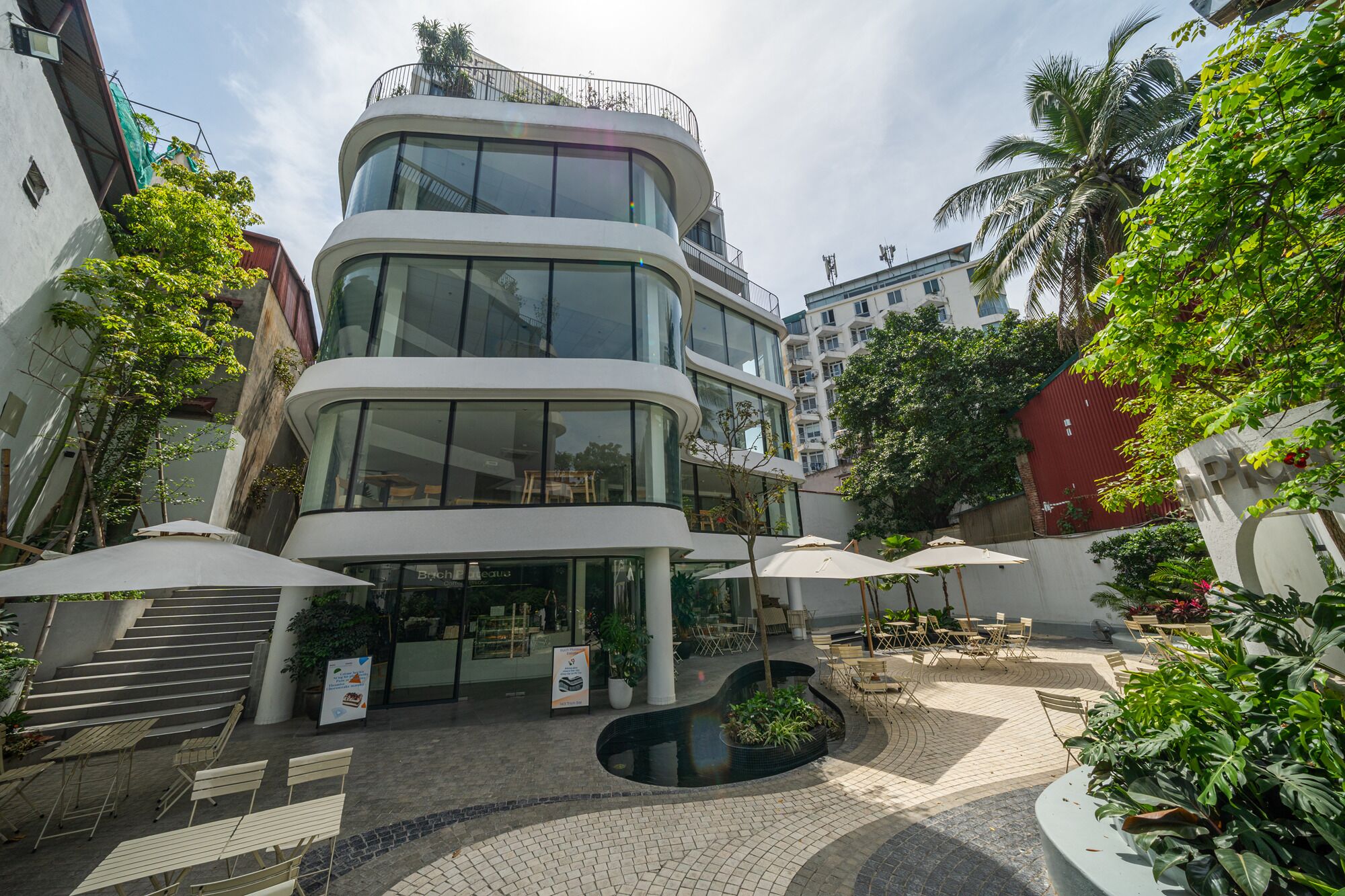 Un edificio moderno con fachada curva, zona de estar al aire libre con sombrillas y una fuente central.