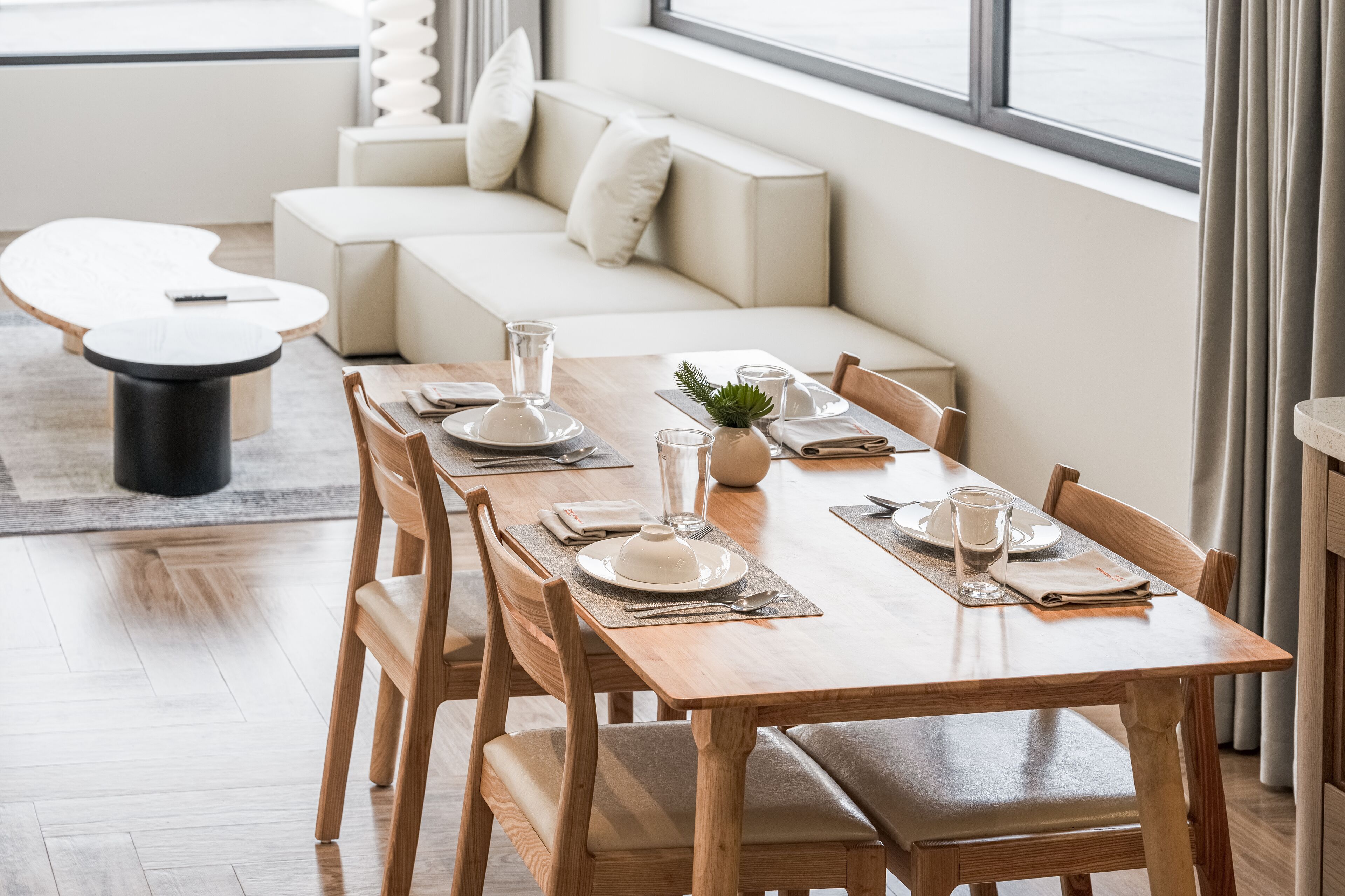 A dining area with a wooden table set for four, surrounded by wooden chairs. A white sofa and a round coffee table are in the background.