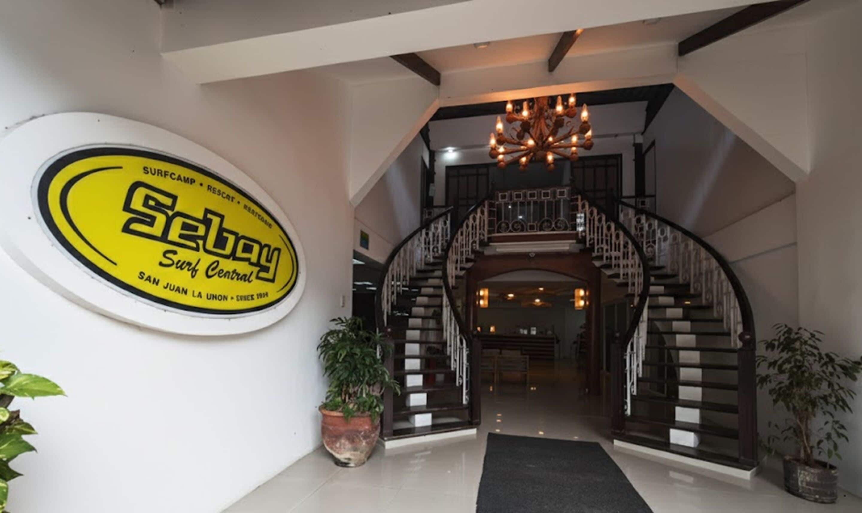 A hotel lobby with a central staircase, a chandelier, and a surfboard logo on the wall.