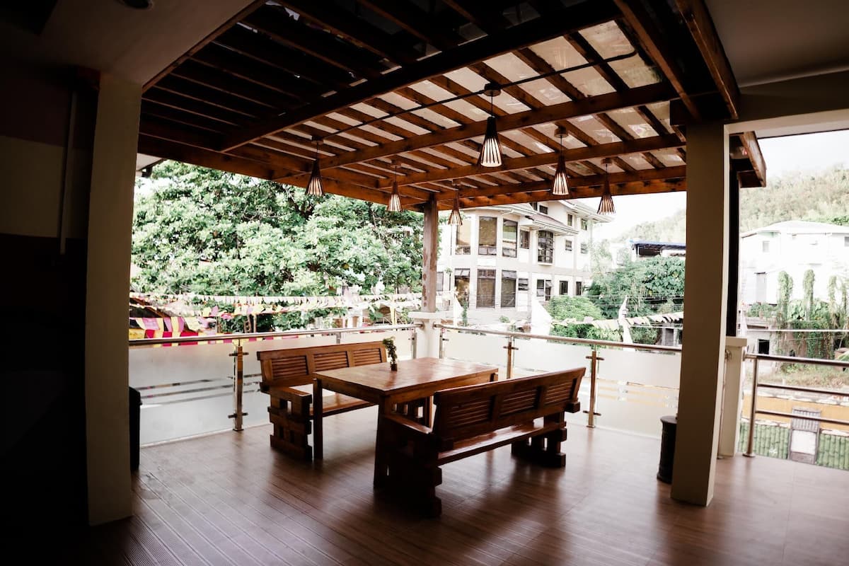 Un área de estar interior con bancos y mesas de madera, techo de madera y vistas a un edificio y árboles.