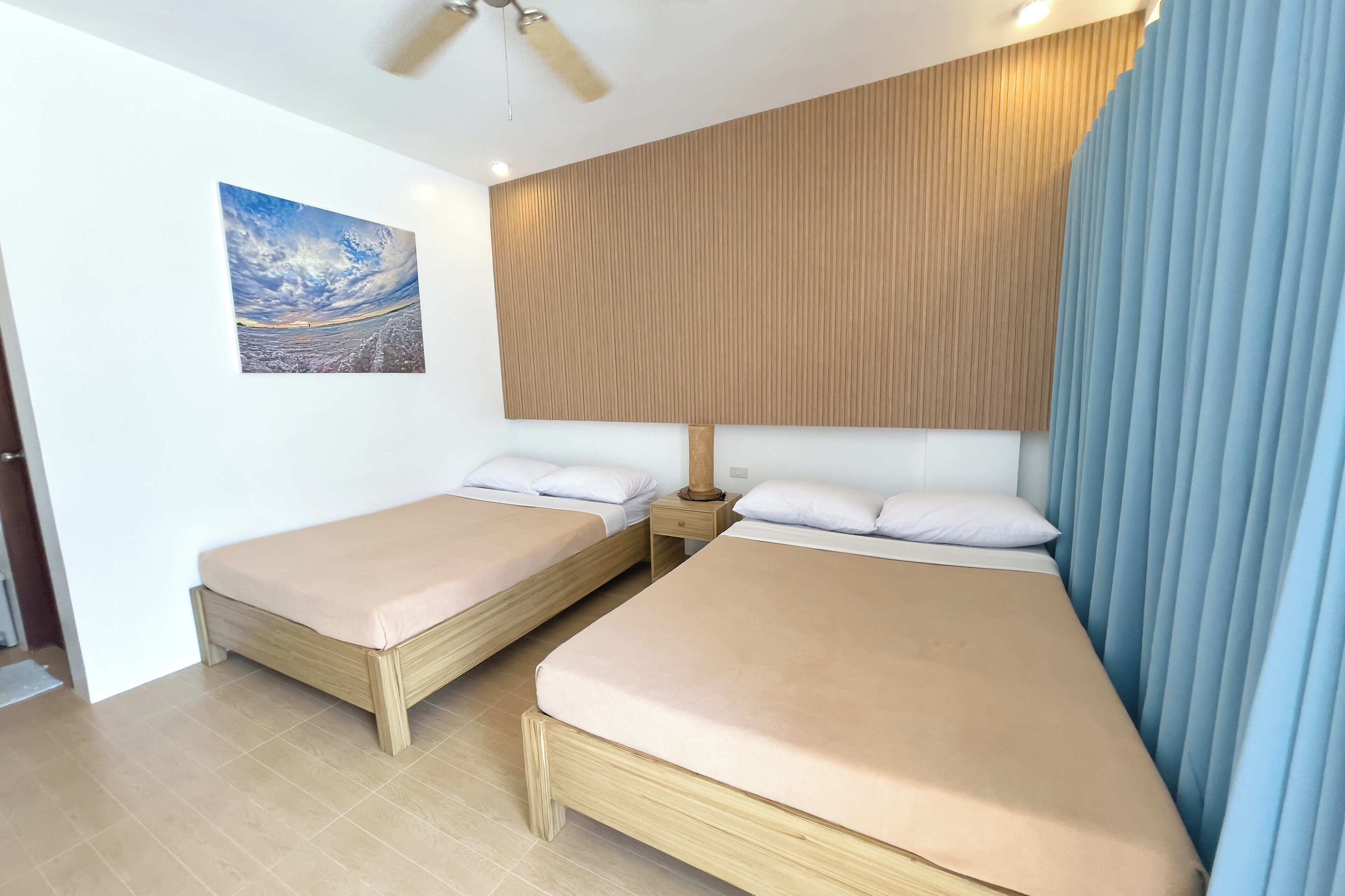 Une chambre avec deux lits simples, un ventilateur de plafond, un mur recouvert de panneaux de bois, une table de chevet et un tableau.