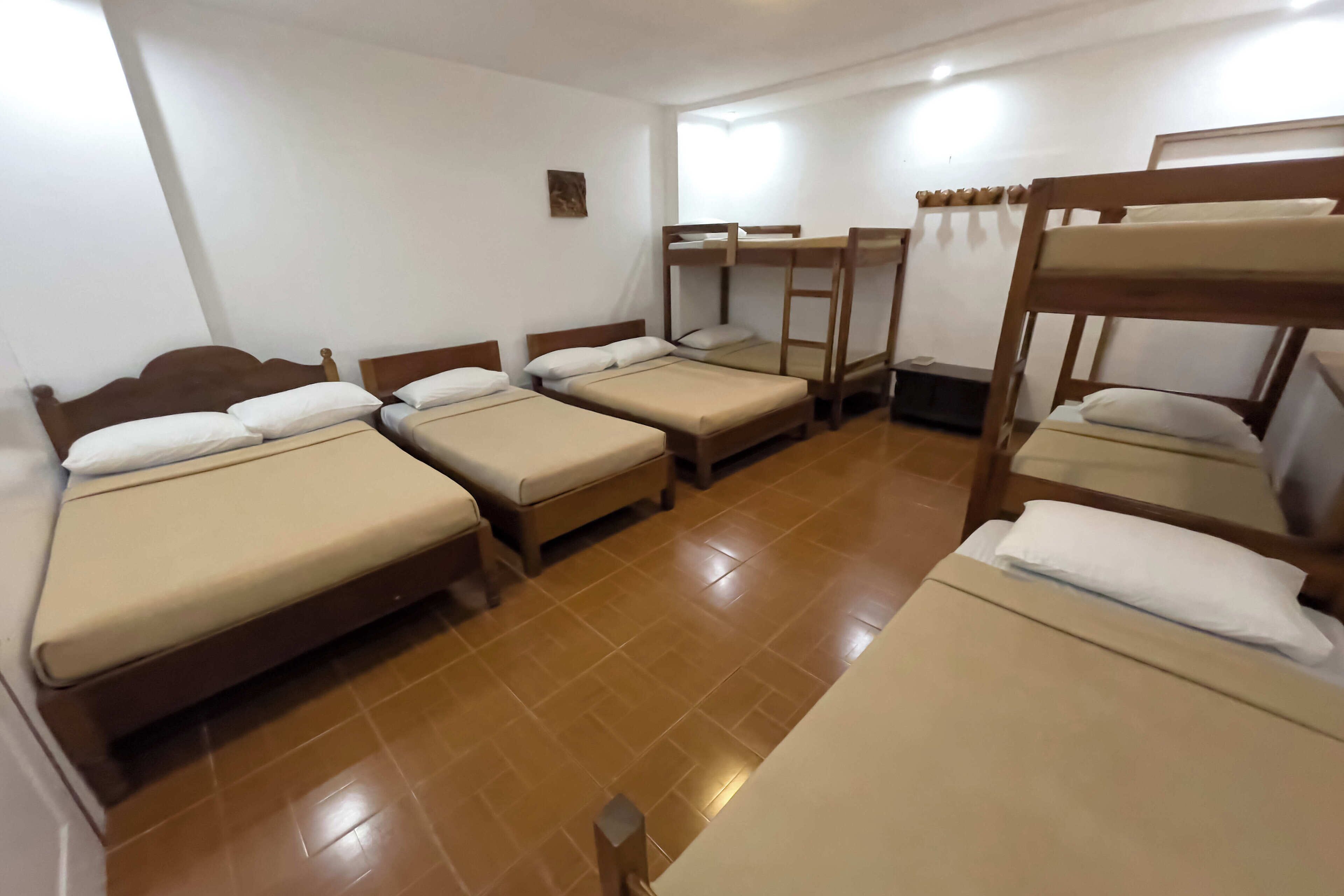 A dormitory room with bunk beds and single beds, wooden headboards, and a tiled floor.