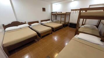 A dormitory room with bunk beds and single beds, wooden headboards, and a tiled floor.