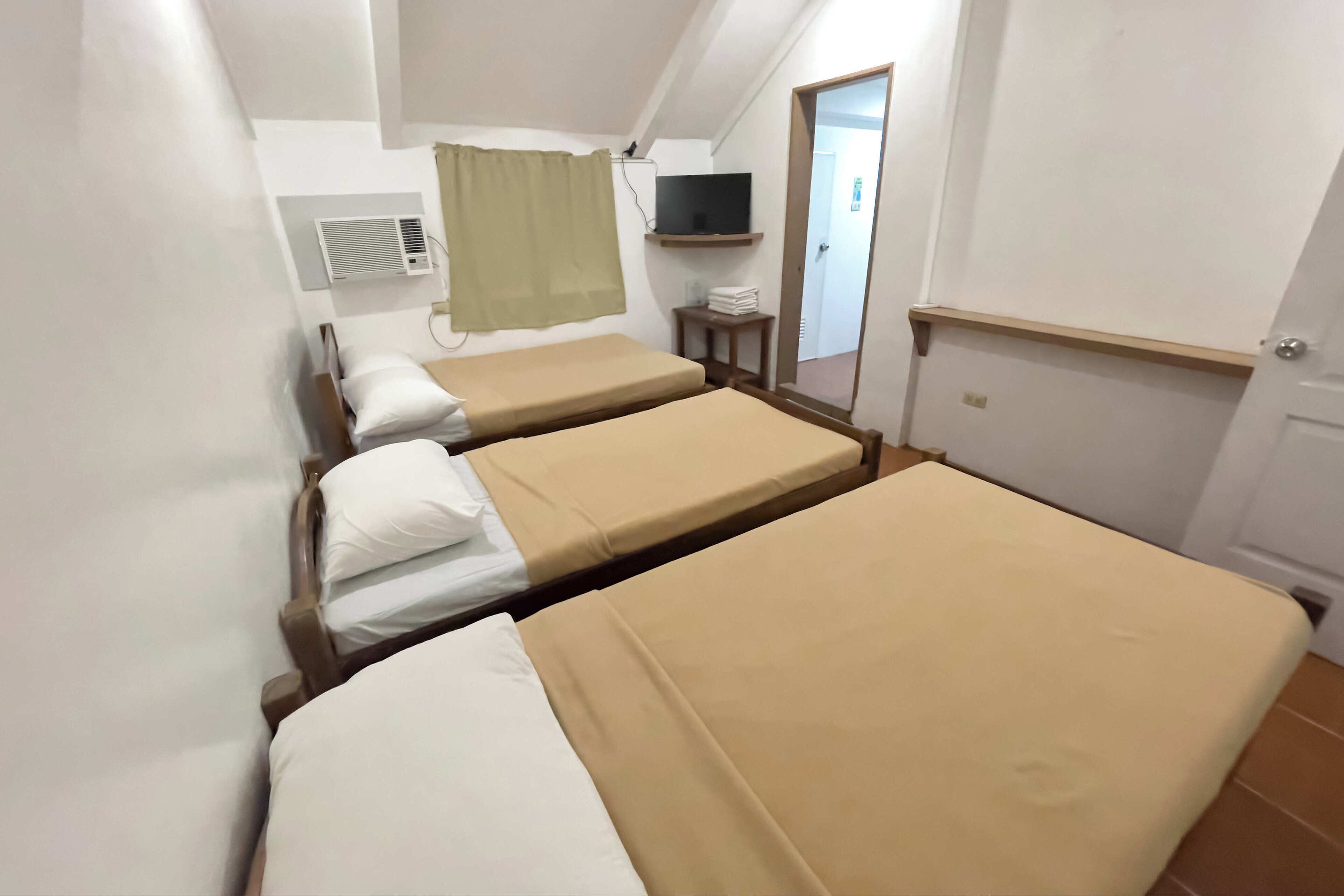 A room with three single beds, a television on a shelf, a small table, and an air conditioning unit.