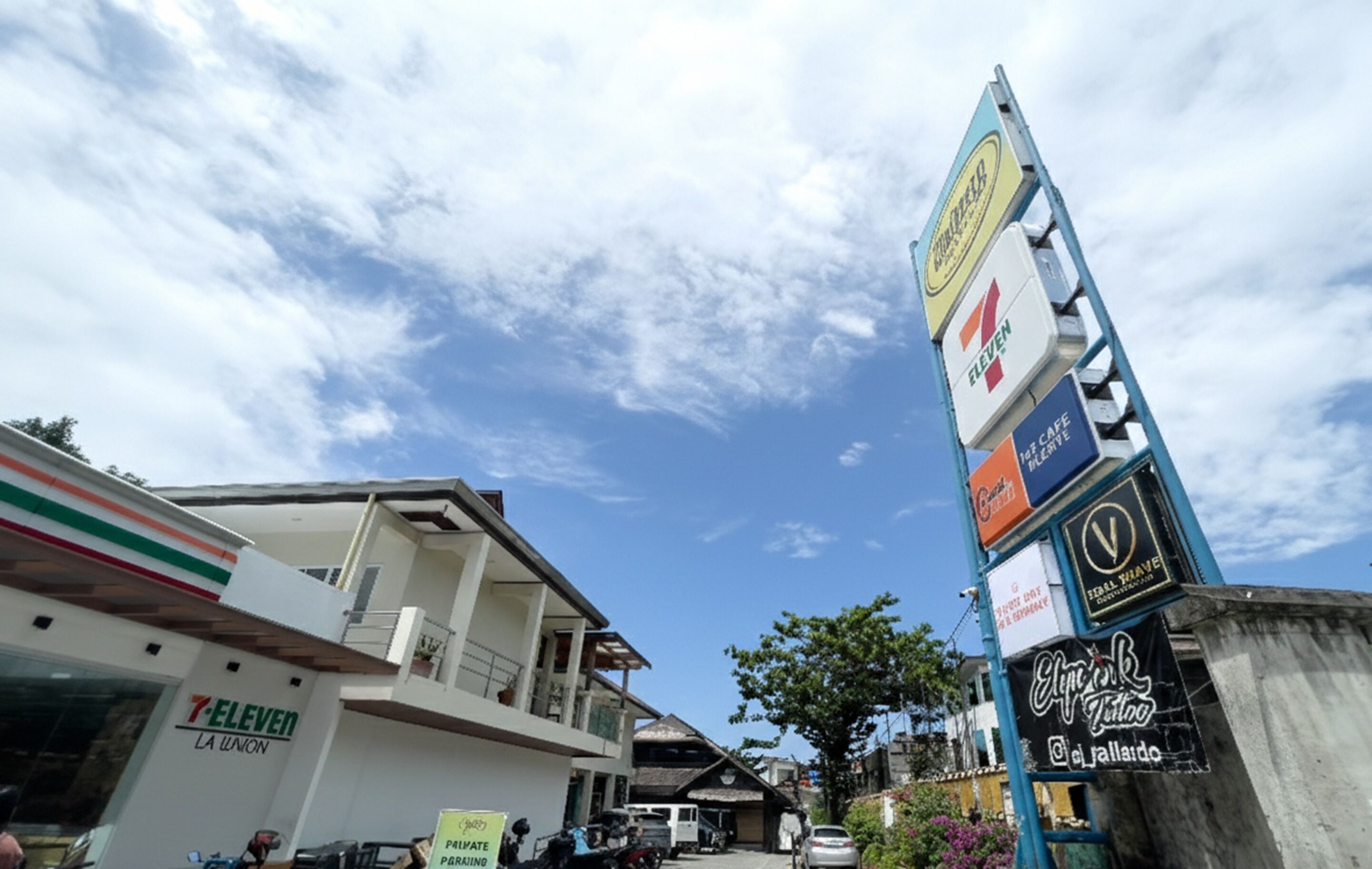 A 7-Eleven store with a signboard advertising various services, a building with balconies, and a clear sky.