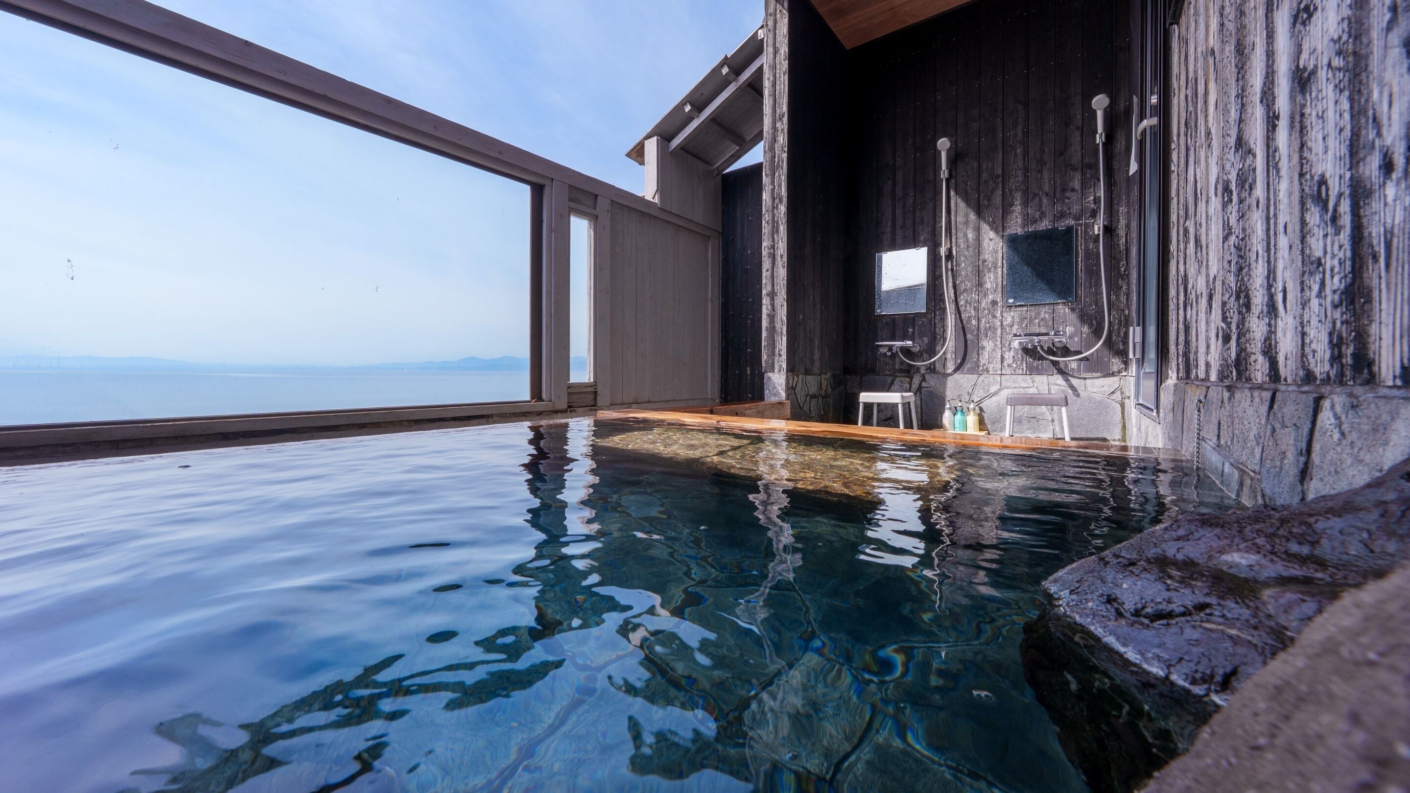 Piscina interior luminosa com vista para o mar, paredes de madeira e uma pequena área de estar.