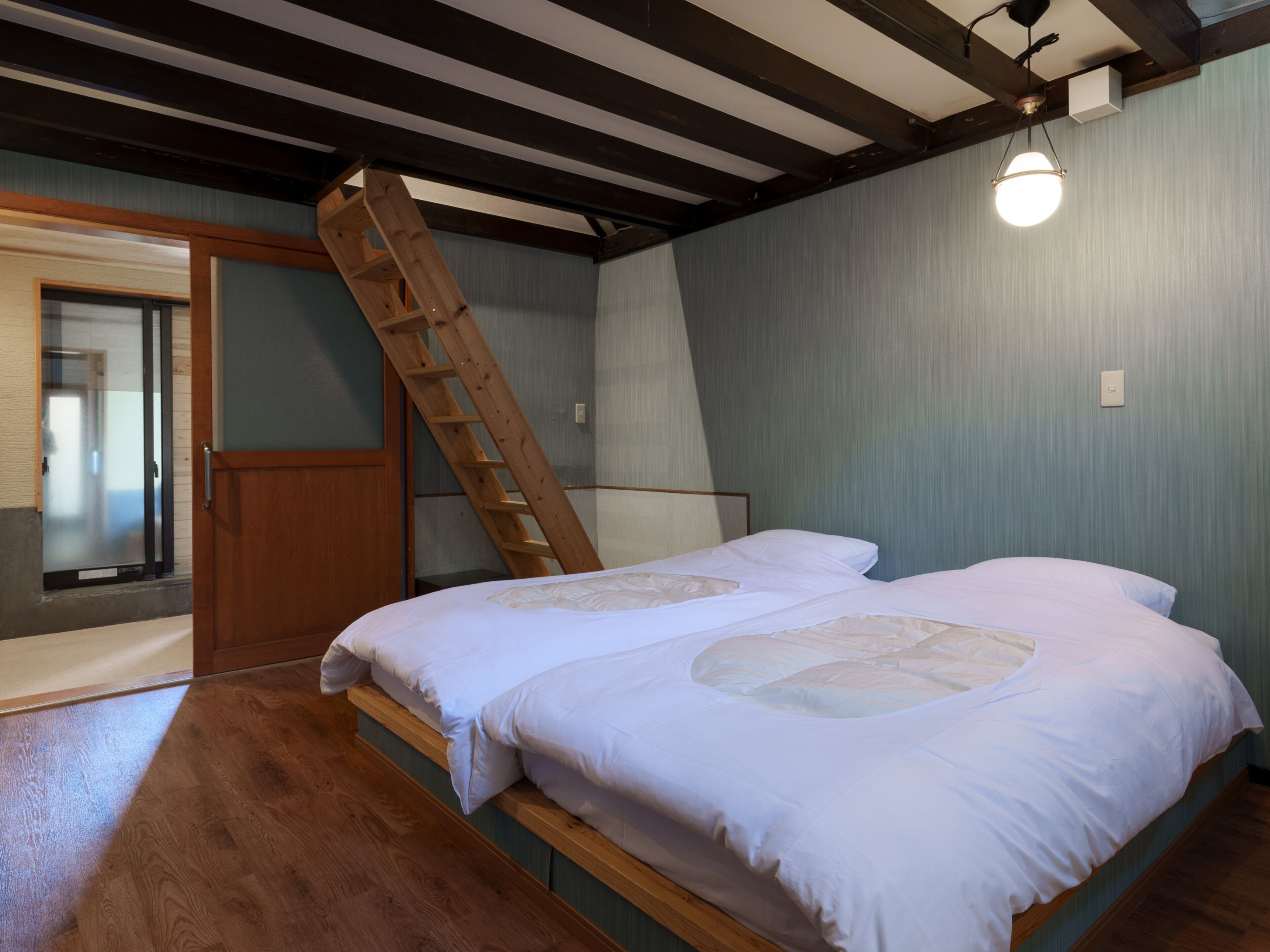 Un dormitorio con una escalera de madera, dos camas con ropa blanca y un suelo de madera, además de una lámpara.