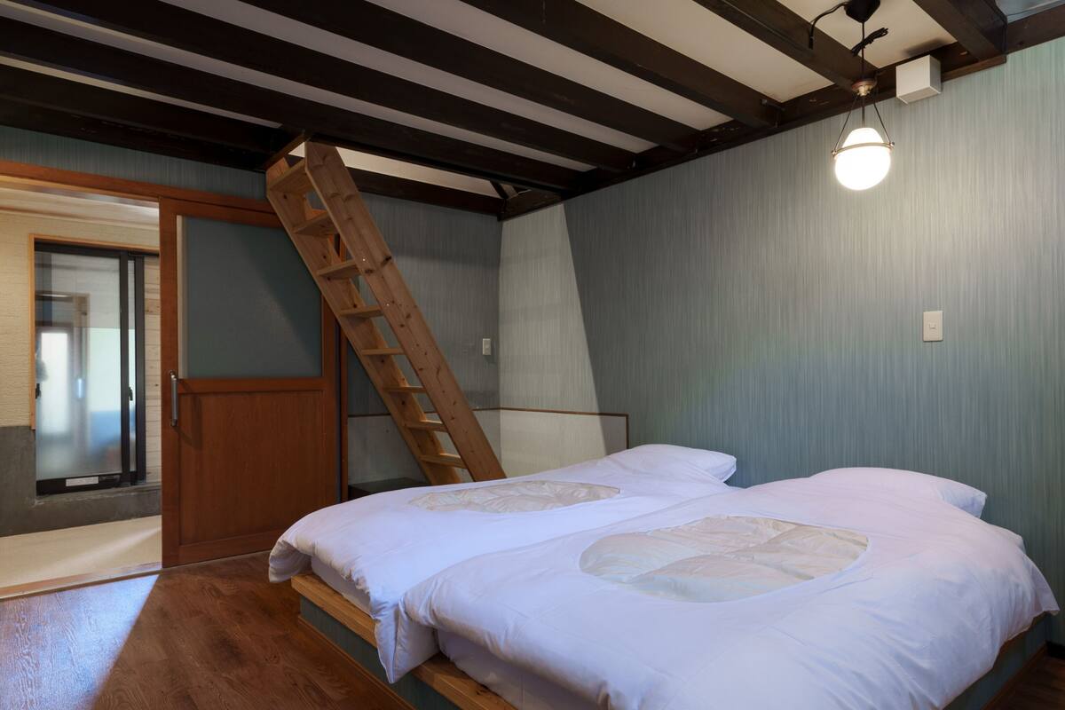 Une chambre avec une échelle en bois, deux lits avec du linge de lit blanc, un plancher en bois et un luminaire.