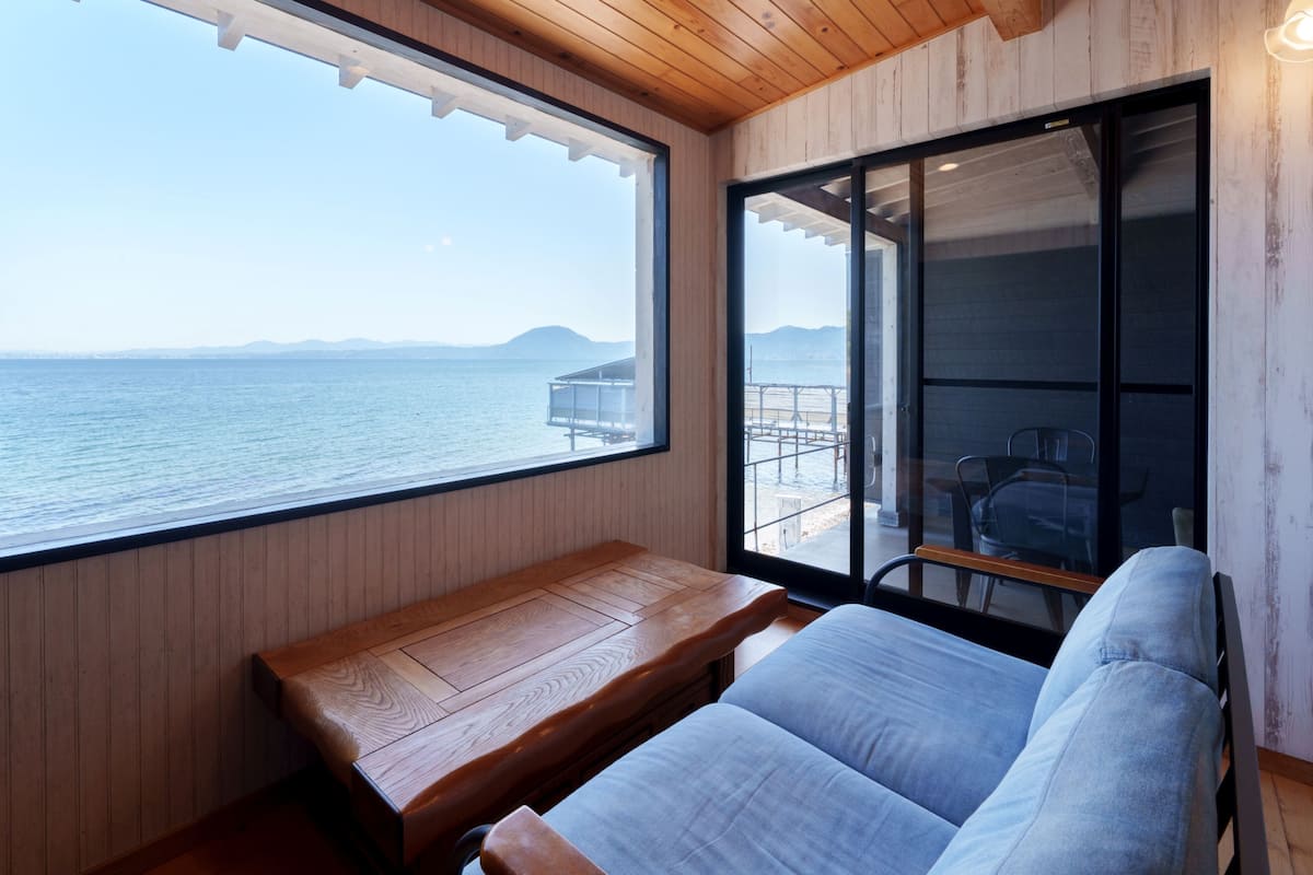 Una habitación con techo de madera, un ventanal con vistas al mar, un banco de madera y un cerramiento de cristal corredera que da a un espacio exterior con una mesa y sillas.