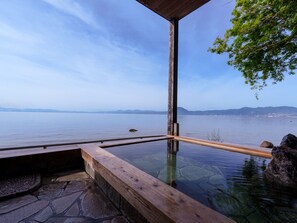 Una terraza de madera con una piscina termal de fondo de cristal, suelo de piedra y vistas a una masa de agua tranquila con tierra a lo lejos.