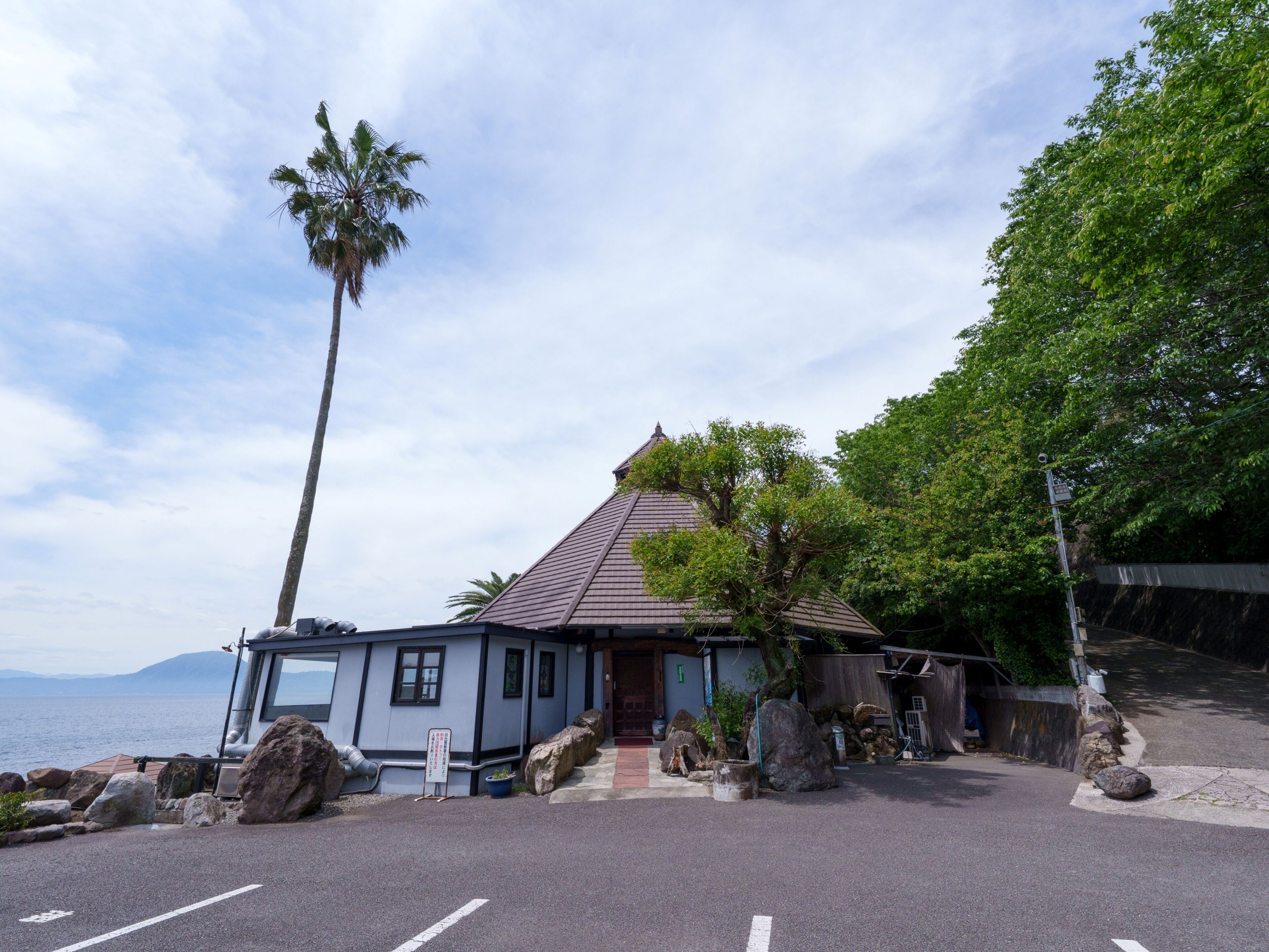 A coastal building with a tiled roof, large rocks, and a palm tree.