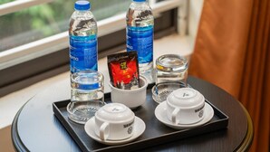 Ein Tablett mit zwei Wasserflaschen, zwei Gläsern, einer Snacks-Packung und zwei Teetassen auf einem Tisch neben einem Fenster.