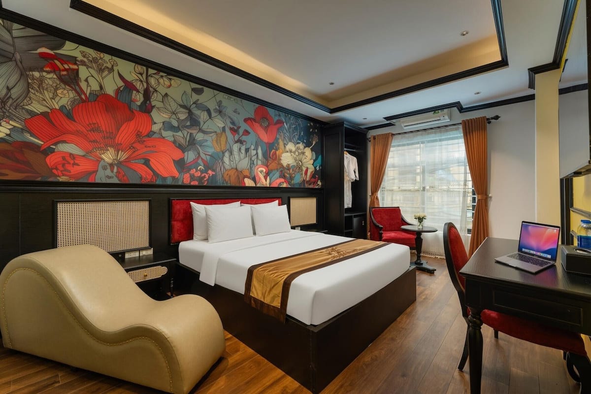 Ein Hotelzimmer mit einem großen Bett, einem Schreibtisch mit Laptop, einem Sessel, einer Couch, Wandbild und einem Fenster mit Vorhängen.