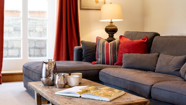 A cozy living room with a grey sofa, a wooden coffee table with books and cups, a lamp, and a window with red curtains. - Stockmans Cottage - Pet Friendly, Todenham (Todenham)