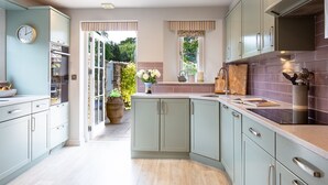 A kitchen with light green cabinets, a white clock, and a view of a garden through an open door. - Old Orchard Cottage - Pet friendly, Moreton-in-Marsh (Moreton-in-Marsh)