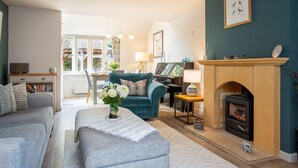 A cozy living room with a fireplace, a piano, and a dining area with a view of the garden. - Old Orchard Cottage - Pet friendly, Moreton-in-Marsh (Moreton-in-Marsh)