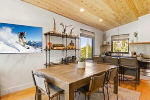 Een ruime eethoek met een houten tafel en stoelen, een groot raam met uitzicht op bomen, en een keuken met moderne apparatuur.