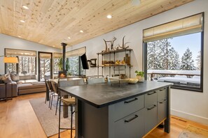 A modern kitchen with a large island, wooden ceiling, and large windows with a snowy view.