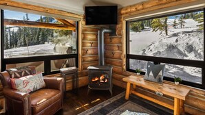 Interior acogedor de una cabaña de troncos con chimenea, sofá de cuero, mesa de madera y ventanas amplias que ofrecen vistas panorámicas de la montaña.