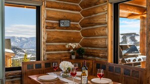 Una mesa de madera para comer con platos blancos y copas de vino, frente a una pared de cabaña de troncos con vistas a las montañas.