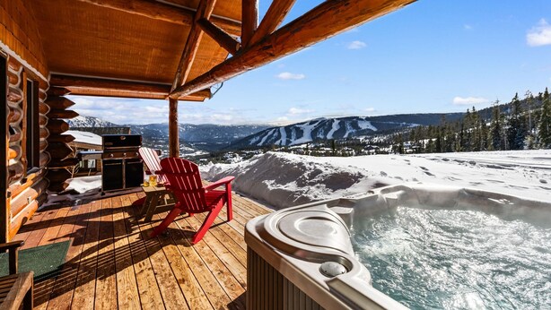 Una terraza de madera con un jacuzzi, sillas rojas y vistas a montañas nevadas.