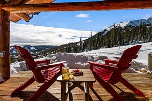 Um deck de madeira com duas cadeiras Adirondack vermelhas, uma mesa com uma garrafa e copos, e um prato de comida.