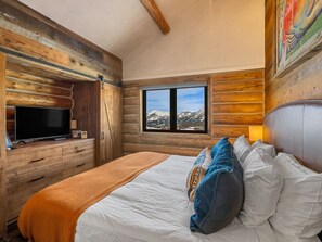 Quarto de cabana de madeira com uma cama, uma TV sobre uma cômoda de madeira e uma janela com vista para as montanhas.