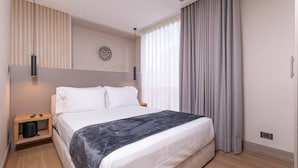 Een moderne hotelkamer met een groot bed, een nachtkastje met een lamp en uitzicht op de stad door grote ramen.