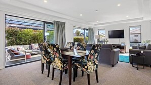 A modern living room with a dining area, large windows, and a flat-screen TV. - Silverstream Kaiapoi Retreat – 3BR Home with Spa & Sun-Filled Living (Kaiapoi)