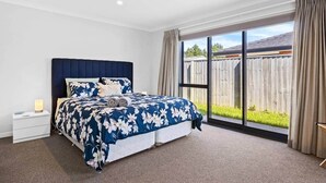 A bedroom with a large bed, a nightstand, a window with curtains, and a view of a backyard. - Silverstream Kaiapoi Retreat – 3BR Home with Spa & Sun-Filled Living (Kaiapoi)