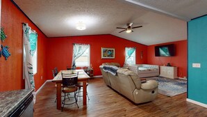 A living room with a red wall, a ceiling fan, a TV, and a dining area with a table and chairs. - Sunset Ave Beach House - Golf Cart Pool Hot Tub (Panama City Beach)