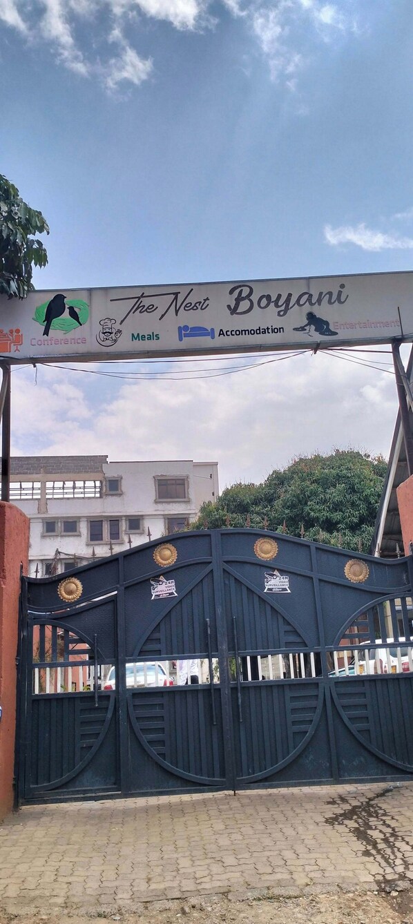 Portão com grade e placa indicando "The Nest Boyani", oferecendo reuniões, refeições, hospedagem e entretenimento.