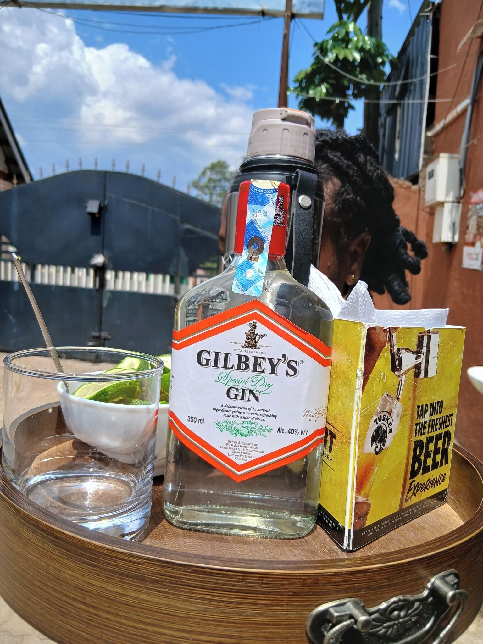 Una botella de ginebra Gilbey's Special Dry, un vaso de agua con lima y una caja de cerveza Tusker sobre una bandeja de madera.